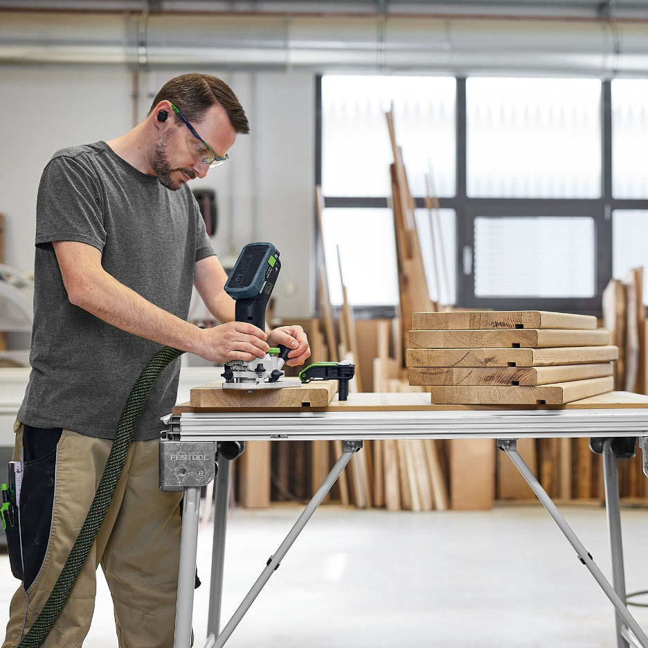 A person wearing safety glasses using the Festool MFKC 700 EB router to trim the edge of a wooden board in a woodworking shop setting