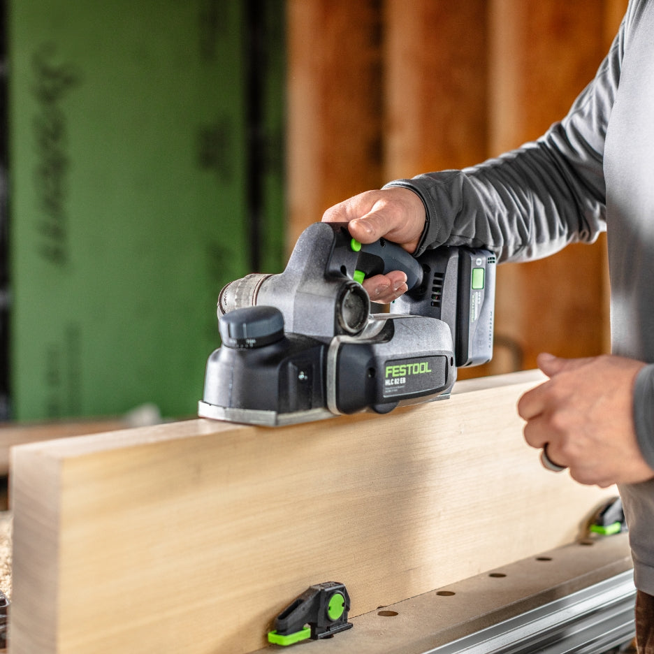 A person uses a Festool HLC 82 EB cordless power planer to smooth the edge of a wooden board in a workshop setting