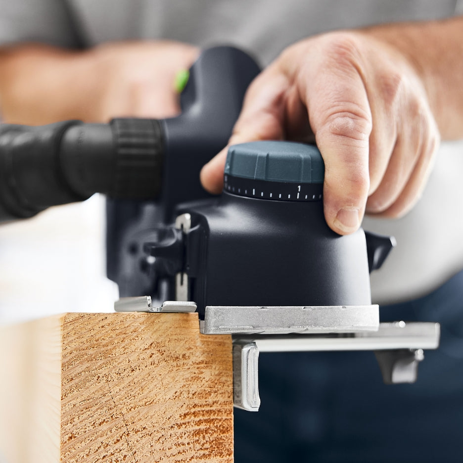 A close-up view of a person using a Festool HLC 82 EB cordless planer to trim the edge of a thick wooden board, highlighting the depth adjustment knob