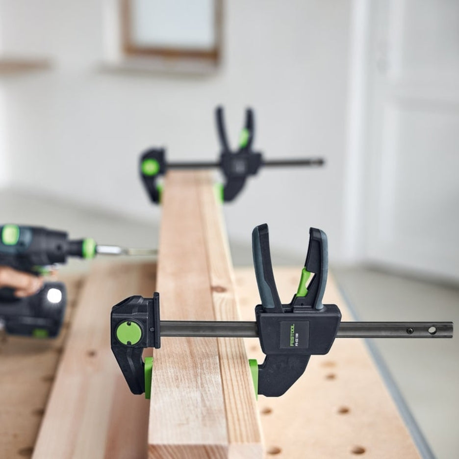 Two Festool One-Handed Clamp FS-EZ 150 holding a piece of wood on a multifunction table with a blurred background.