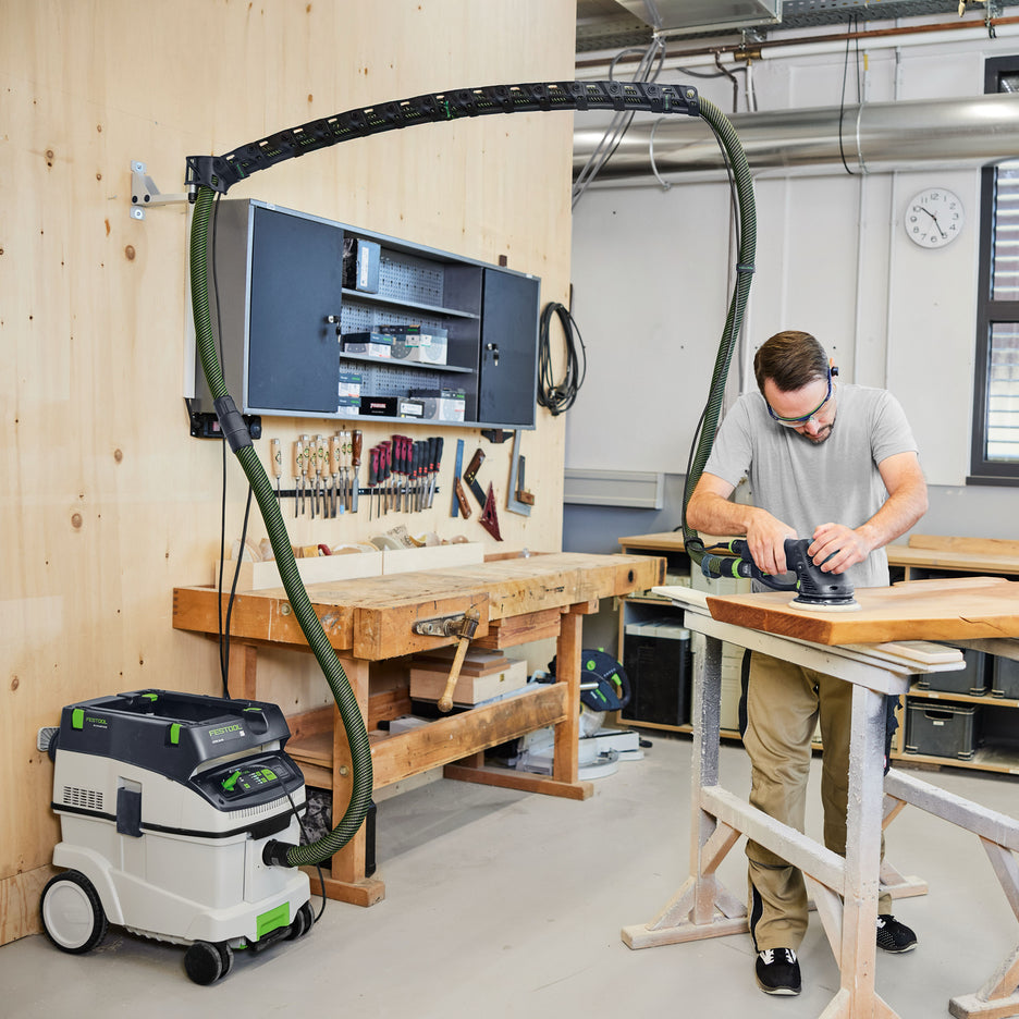 Festool wall-mount boom arm supporting hose and Plug-it power cord overhead while man sands wood slab on sawhorses