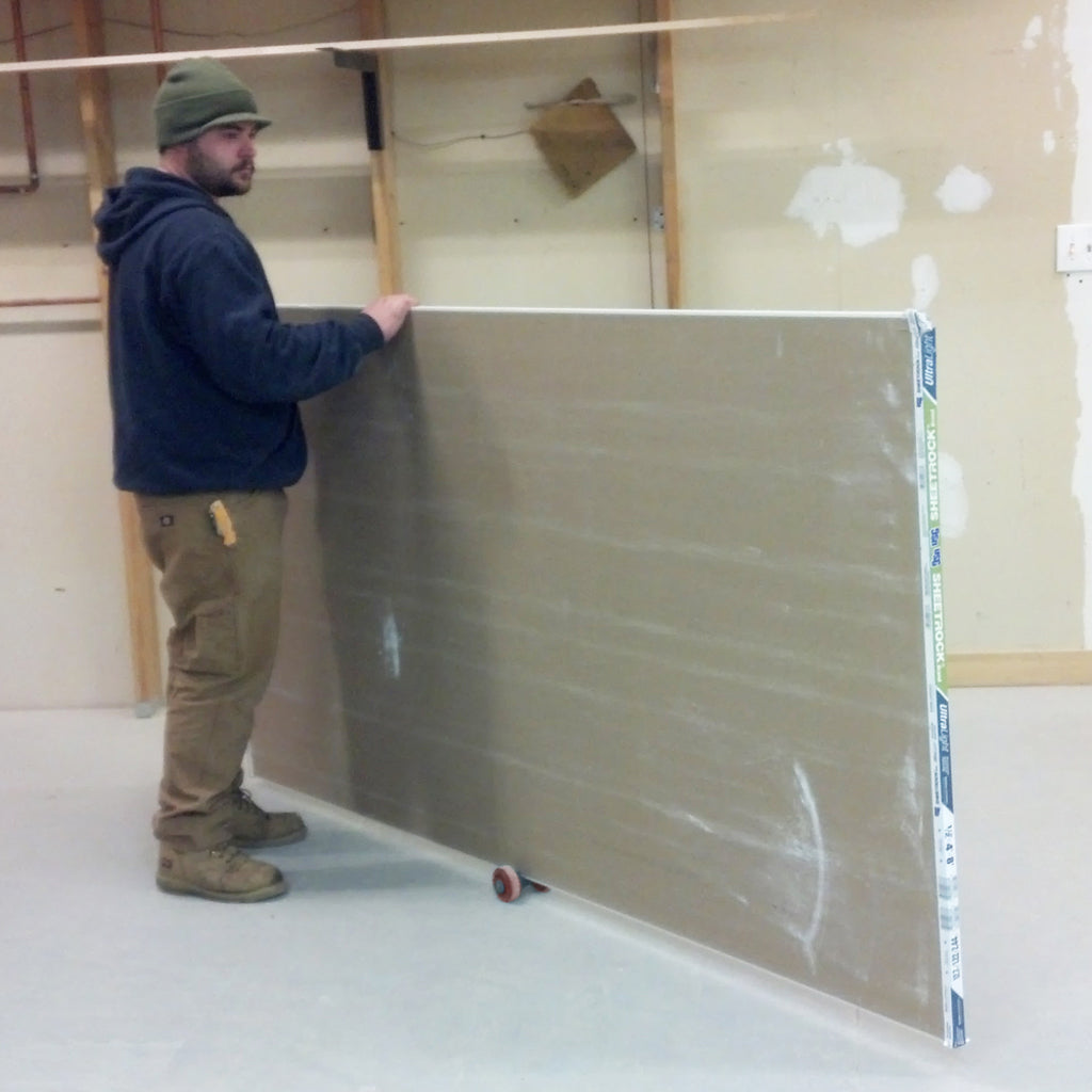 A worker moves a full sheet of drywall by himself, balanced on the Speed Skate.