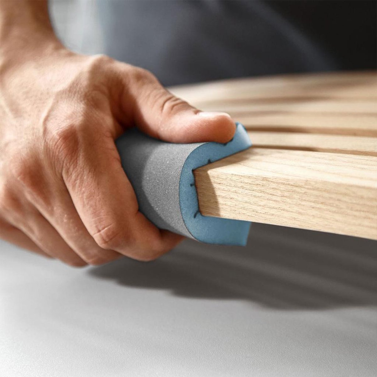 A bare hand holds an abrasive sponge folded over a narrow wood component to sand three faces simultaneously.