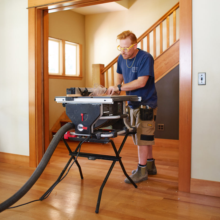Woodworker rips a narrow piece of solid wood trim on SawStop Compact Saw on folding stand with dust collection inside house.