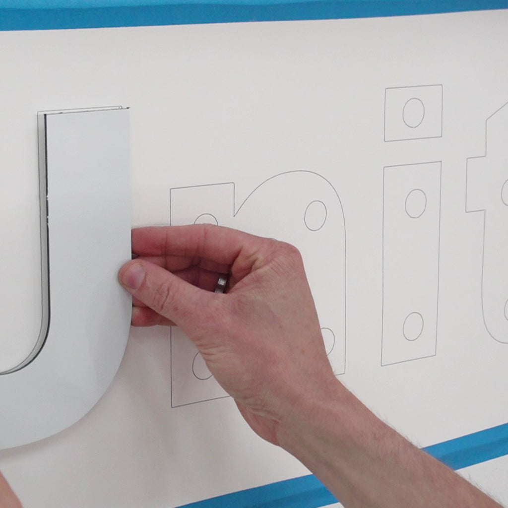 A maker positions a letter cutout precisely according to the drawn layout for a sign, created with CNC stylus and pen.