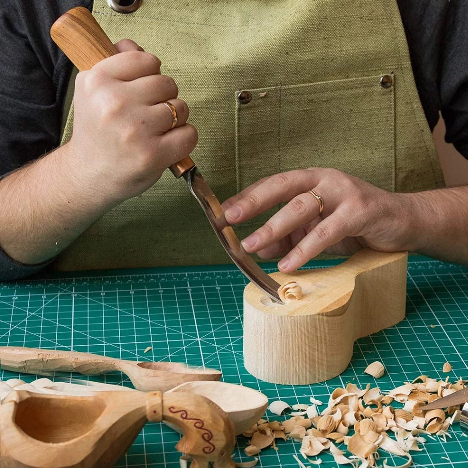 Person carving wooden spoons with a BeaverCraft chisel on a green cutting mat.