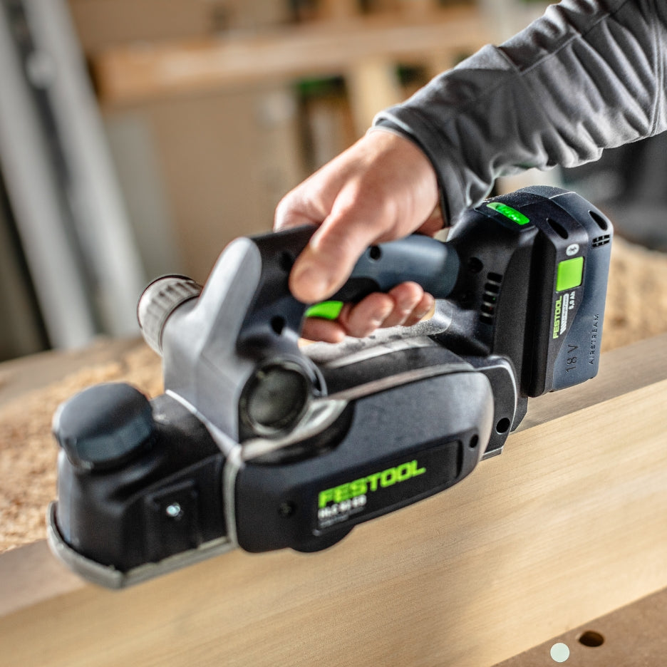 A close-up side view of a Festool HLC 82 EB cordless power planer held by a person as it smooths the top surface of a wooden board
