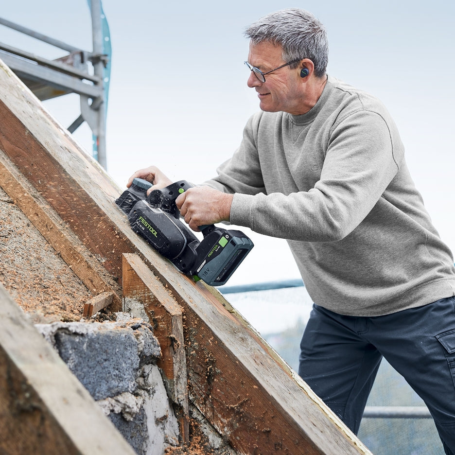 A man uses a Festool HLC 82 EB cordless planer to smooth the edge of a timber roof rafter on a construction site