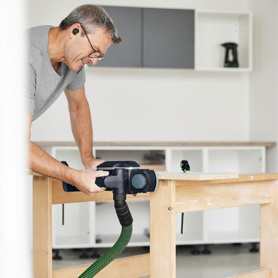 A person in a workshop uses a Festool HLC 82 EB cordless planer connected to a dust extraction hose to smooth the edge of a wooden board