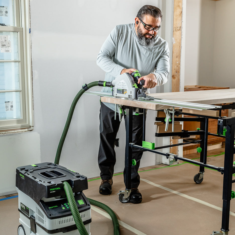A man wearing safety glasses and a light gray long-sleeve shirt operates a Festool TSC 55 KS cordless track saw with guide rail and CTC MIDI I Cordless Dust Extractor to cut a hardwood counter top on STM 1800 cutting table in a house being renovated.