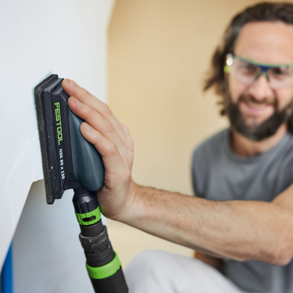 Man sands drywall near corner with Festool hand sanding block connected to dust extraction hose.