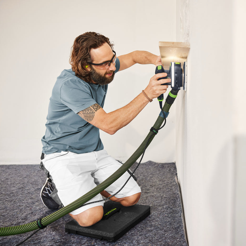 Man kneels on a Festool Seat Cushion on the ground as he sands the wall underneath a shelf.