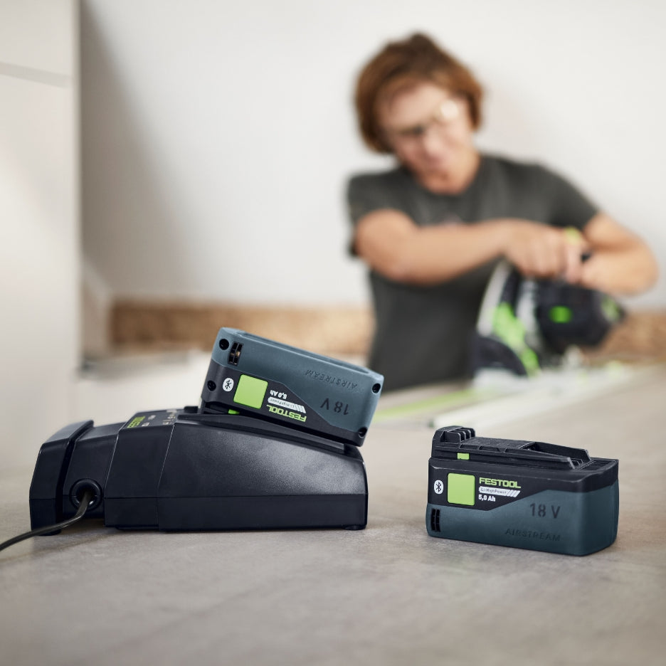 Pentool battery and charger on a table with a blurred person using a power tool in the background.